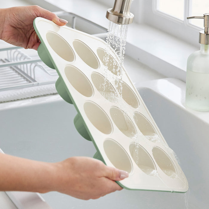 GreenLife sage green ceramic nonstick muffin pan being rinsed under a kitchen faucet in a bright kitchen.