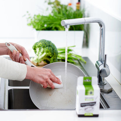 Hand cleaning a skillet with GreenPan accessory kit under running water, vegetables and soap visible nearby.