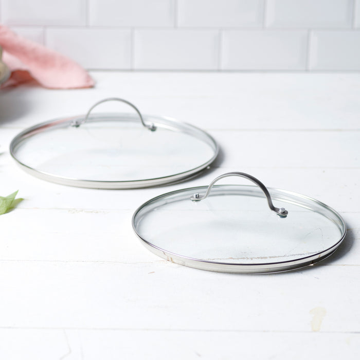 Two glass cookware lids on a white surface with a tiled wall background