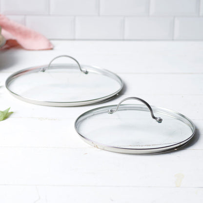 Two glass cookware lids on a white surface with a tiled wall background