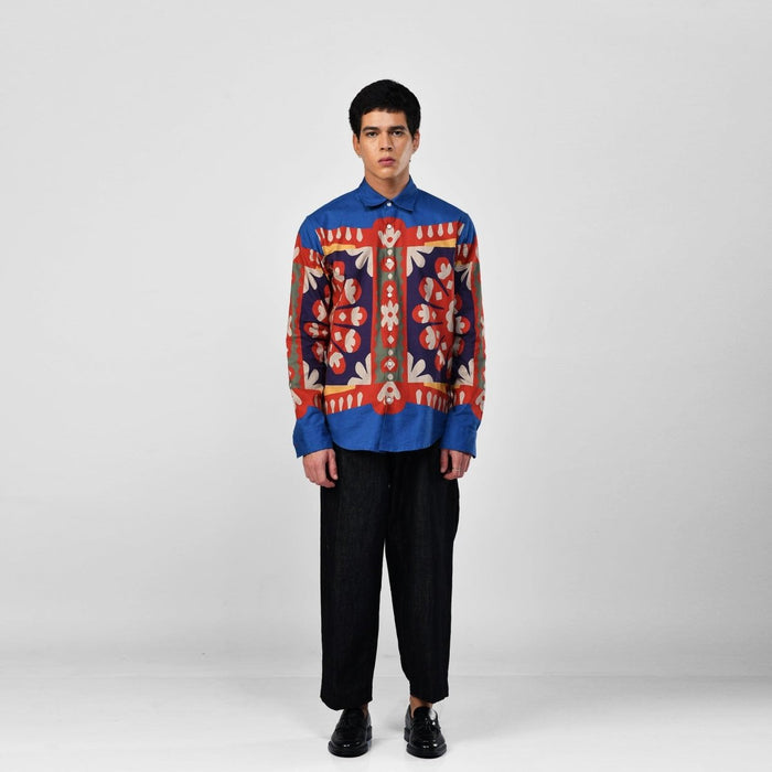 Man wearing the blue Kanaat shirt with black trousers, full length front view, red, navy and orange pattern panel