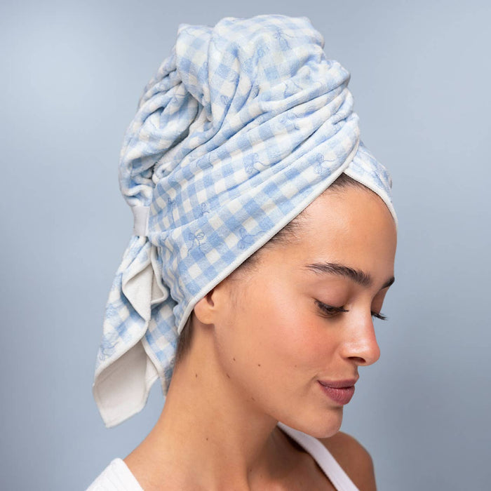 A woman wearing a light blue and white gingham checkered cotton towel wrapped around her head, shown in profile against a soft light blue-gray background. Microfibre Hair Towel | Blue Gingham Hair Wrap | Kiyo Beauty