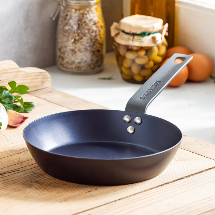 Carbon steel 8-inch frypan on a wooden counter, with jars, eggs, and olives in the background.