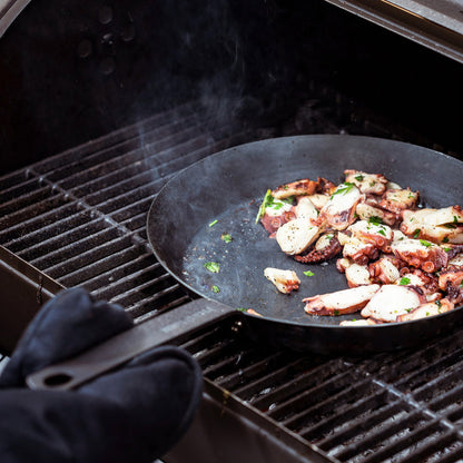 Carbon steel 8-inch frypan with cooked octopus and herbs on a grill, held by a black oven mitt.