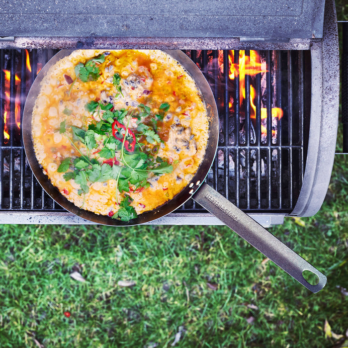 Carbon steel frypan with stew, herbs, and chili on a grill over open flames, grassy ground visible below.