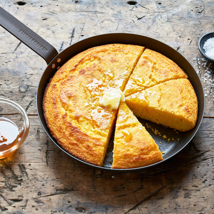 Cornbread topped with butter and honey in an 8-inch Merten  Storck carbon steel frypan on a wooden table.