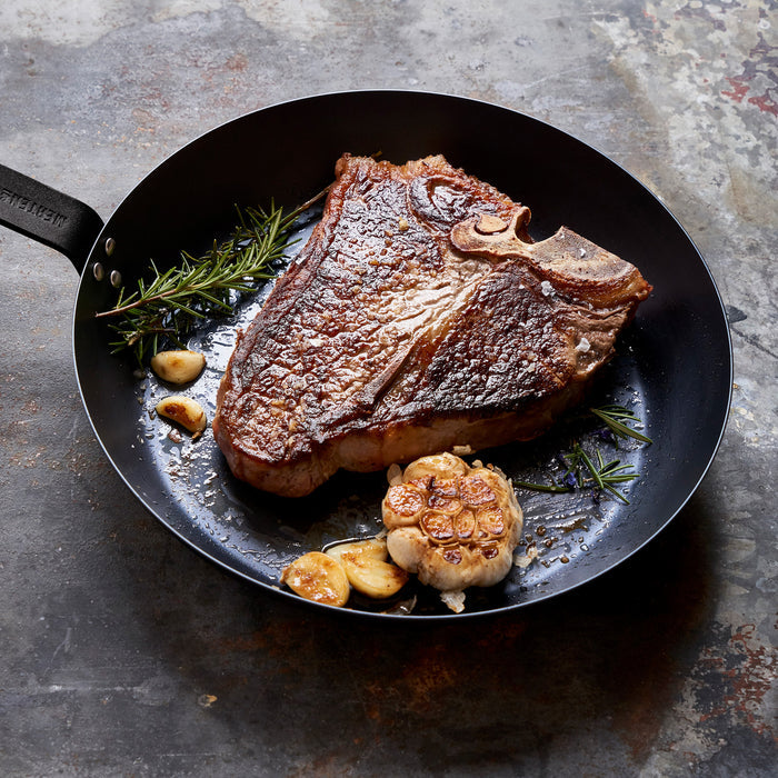 Merten  Storck carbon steel frypan with a cooked T-bone steak, rosemary, and garlic on a rustic surface.