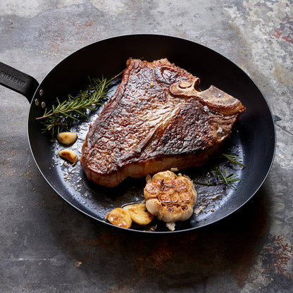 Merten  Storck carbon steel frypan with a cooked T-bone steak, rosemary, and garlic on a rustic surface.