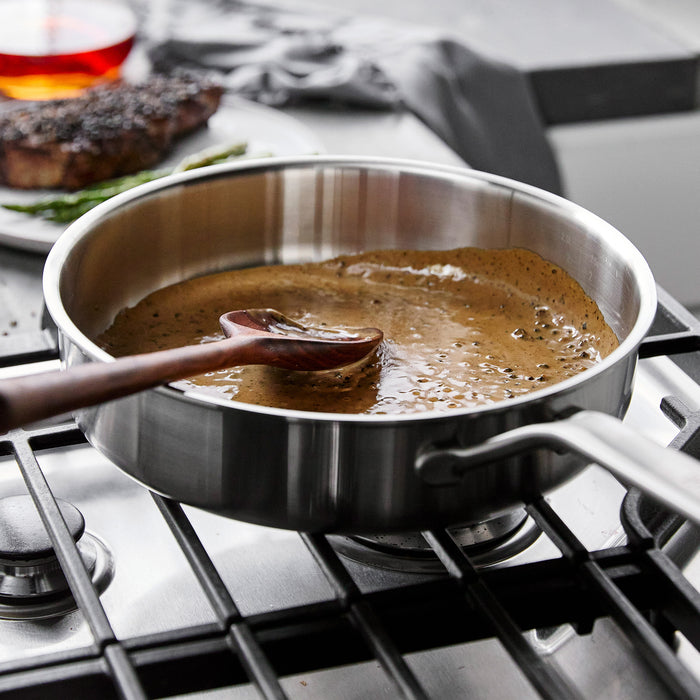 Stainless steel sauté pan with sauce being stirred by a wooden spoon on stove, steak visible in background.