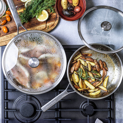 Stainless steel sauté pan with lid on stovetop, potatoes and herbs cooking nearby.