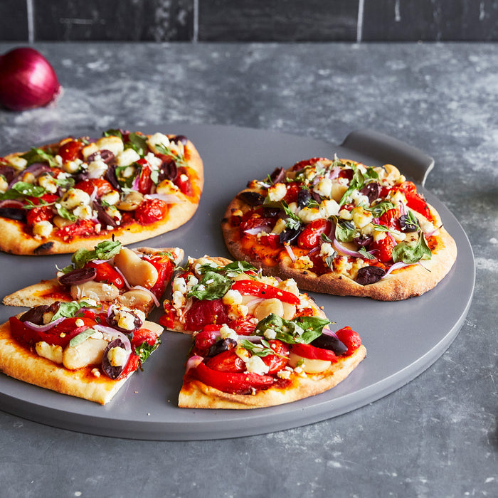 Three mini veggie pizzas on a gray Premiere Ovenware 16.5 round pizza pan, one slice pulled out on a kitchen counter.