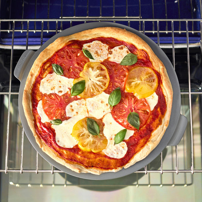 Homemade pizza being placed onto a gray round pizza baker tray in a kitchen oven.
