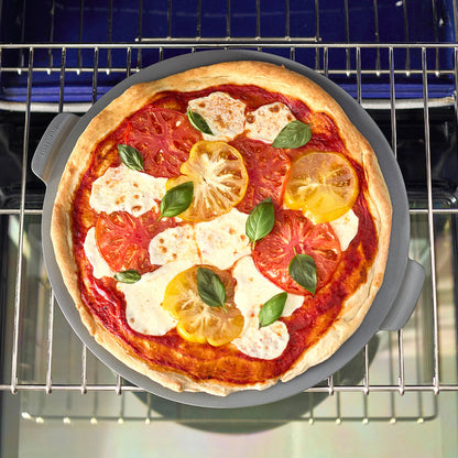 Homemade pizza being placed onto a gray round pizza baker tray in a kitchen oven.