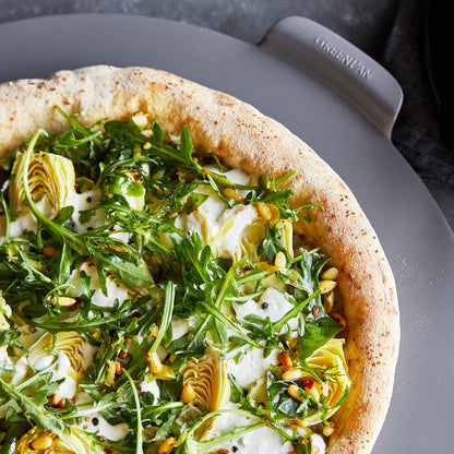 Pizza topped with arugula, artichoke hearts, pine nuts, and cheese on a gray 16.5-inch round pizza baker.