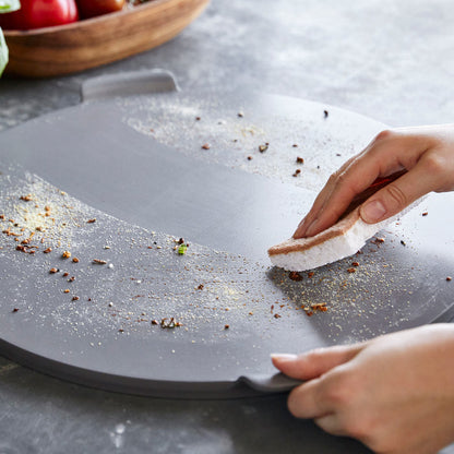 A hand sponging crumbs off a gray Premiere Ovenware 16.5 round pizza baker on a kitchen countertop.