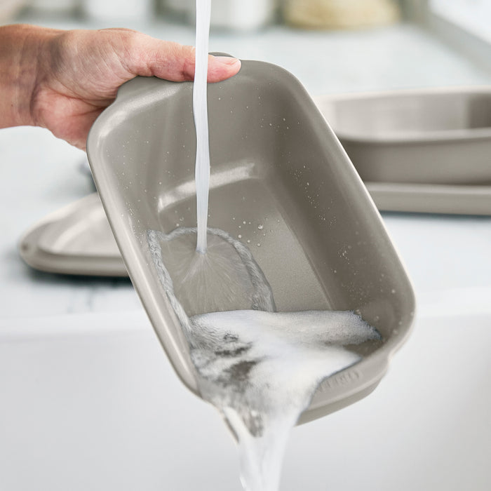 Ceramic nonstick taupe loaf pan being rinsed under running water at a kitchen sink.