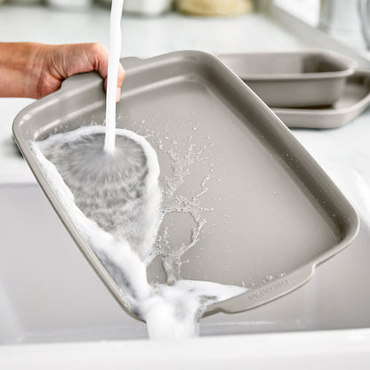 Hand washing a ceramic nonstick 13x9 quarter sheet pan set in a kitchen sink.