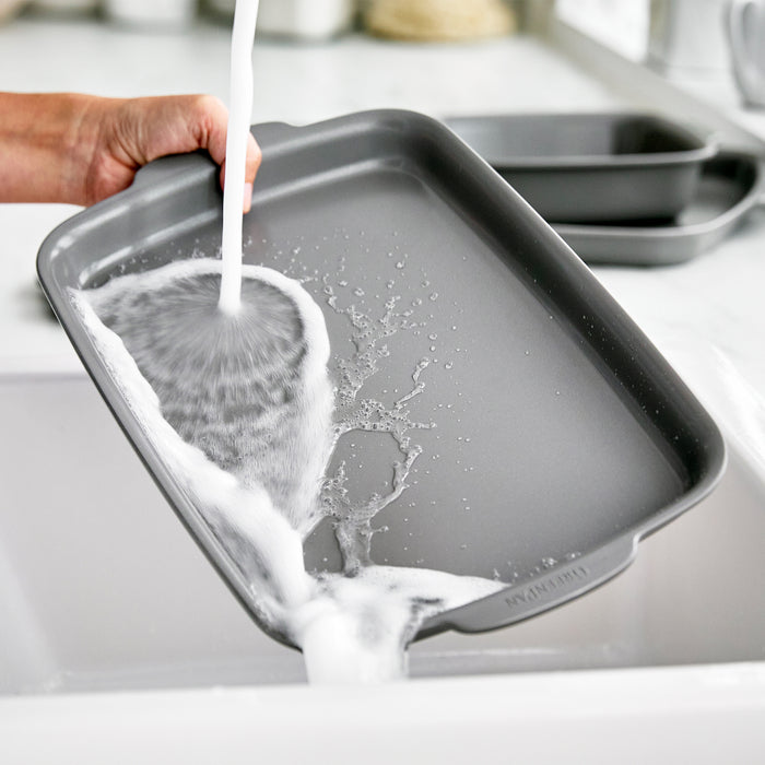 Hand washing two silver ceramic quarter sheet pans under running water in a kitchen sink.
