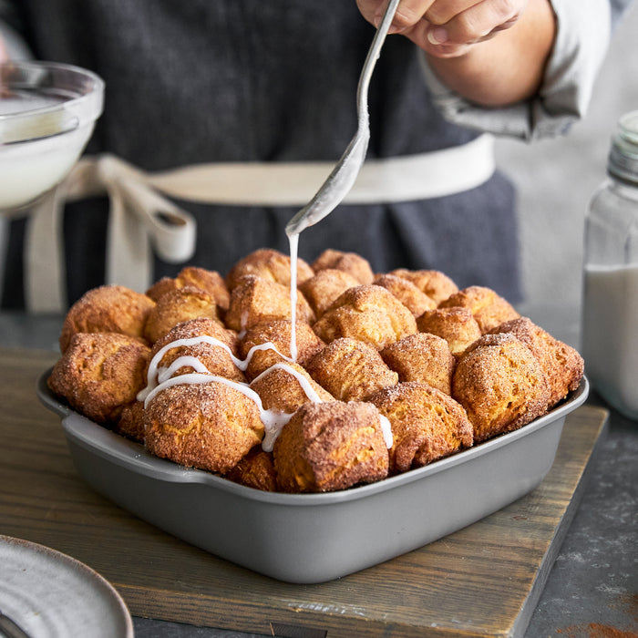 Icing being drizzled over monkey bread in a ceramic nonstick cake pan set on a wooden surface.