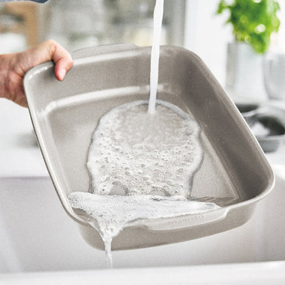 Hand rinsing soapy taupe ceramic nonstick cake pans under running water in a bright kitchen sink.