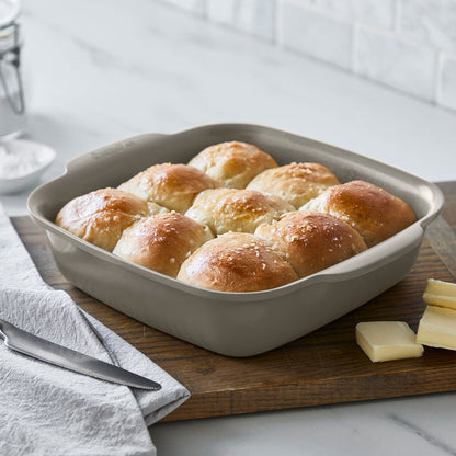 Eight-inch square ceramic baker with built-in handles holding nine golden brown dinner rolls.