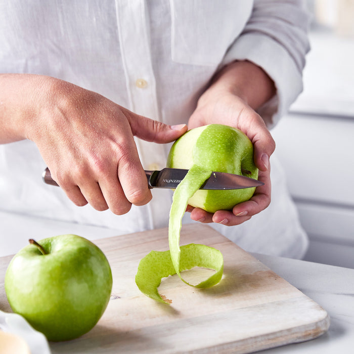 Hand peeling a green apple with a Premiere Titanium Cutlery knife; second apple on wooden cutting board.