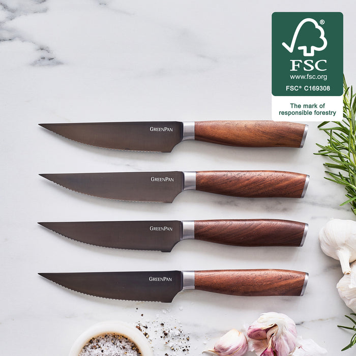 Four titanium steak knives with walnut handles on a white surface, arranged with sprigs of herbs and garlic cloves nearby.