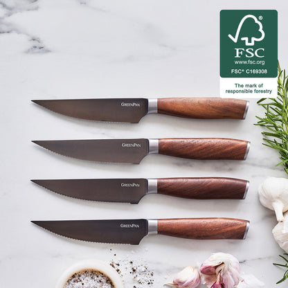 Four titanium steak knives with walnut handles on a white surface, arranged with sprigs of herbs and garlic cloves nearby.