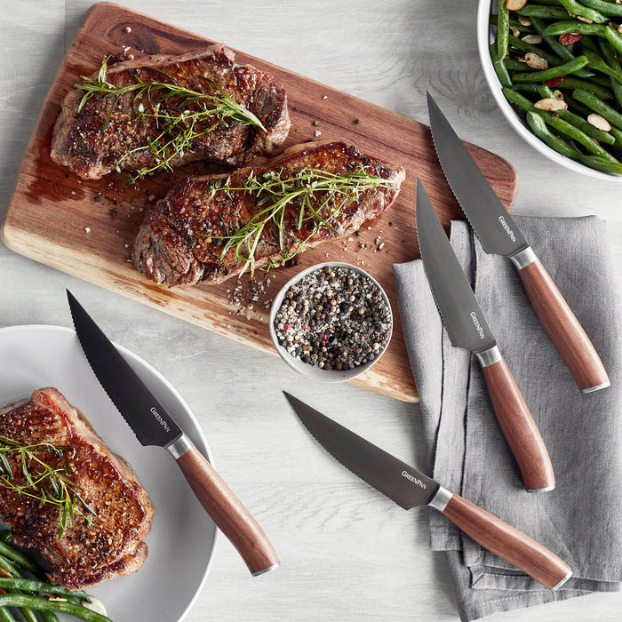 Two cooked steaks with herbs and vegetables next to a set of four titanium steak knives.