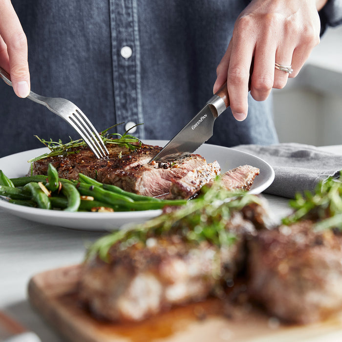 A steak and green beans being cut with a stainless steel steak knife; another steak rests on a wooden board nearby.