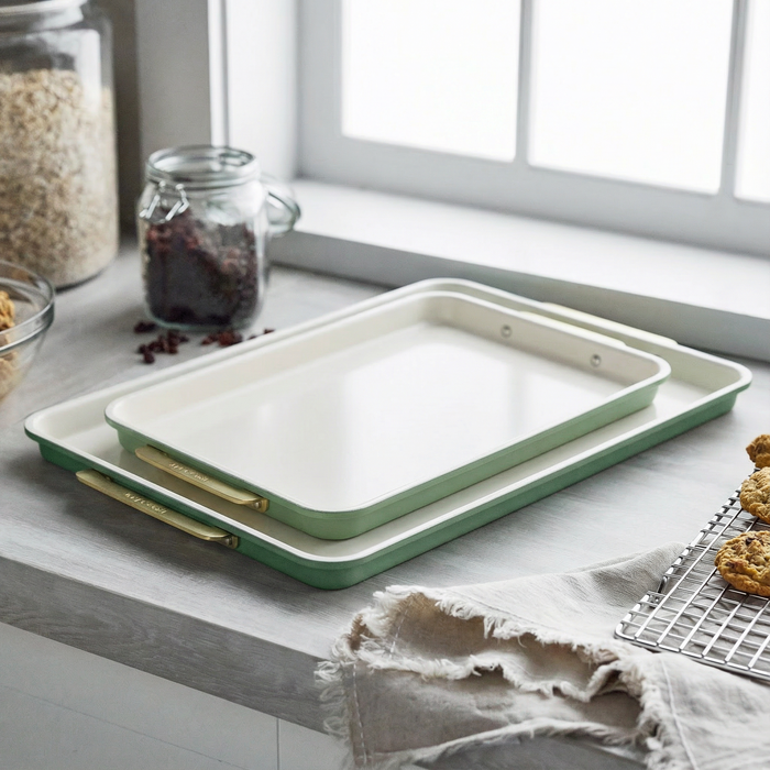 Two sage green sheet pans from a 2-piece bakeware set rest on a kitchen counter beside cookies cooling by the window.