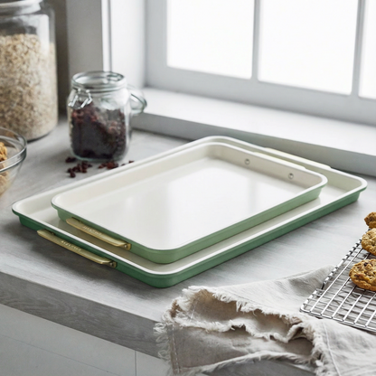Two sage green sheet pans from a 2-piece bakeware set rest on a kitchen counter beside cookies cooling by the window.