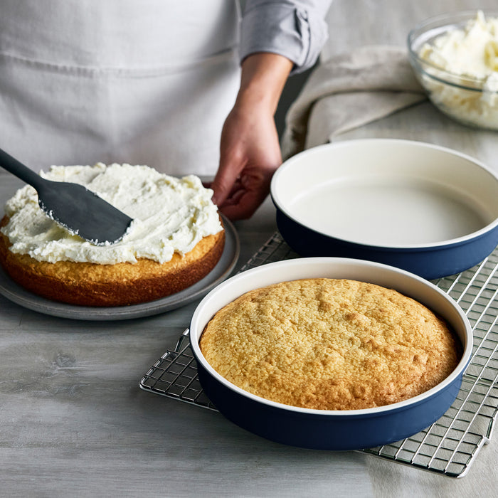 Person frosting a cake next to another layer in a navy Reserve Bakeware pan from an 8-piece set on a cooling rack.