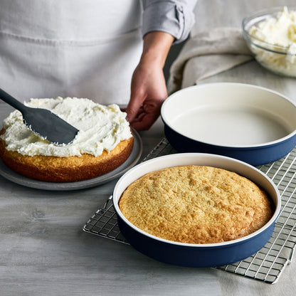 Person frosting a cake next to another layer in a navy Reserve Bakeware pan from an 8-piece set on a cooling rack.