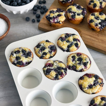 Blueberry muffins in an 8-piece Reserve Twilight bakeware set, with loose blueberries and sugar beside the pans.