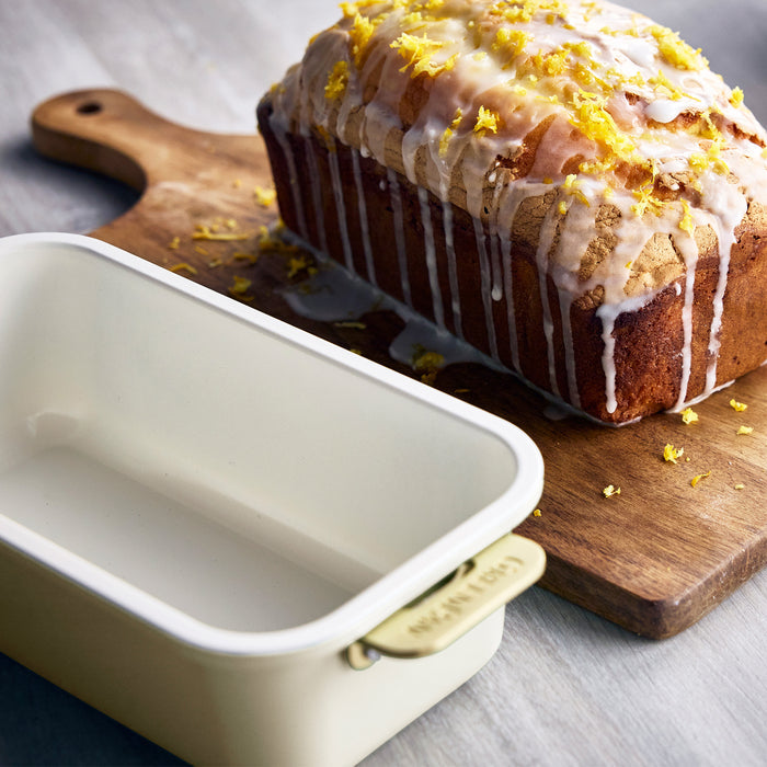 Loaf of cake with lemon drizzle on a wooden cutting board next to a cream loaf pan.