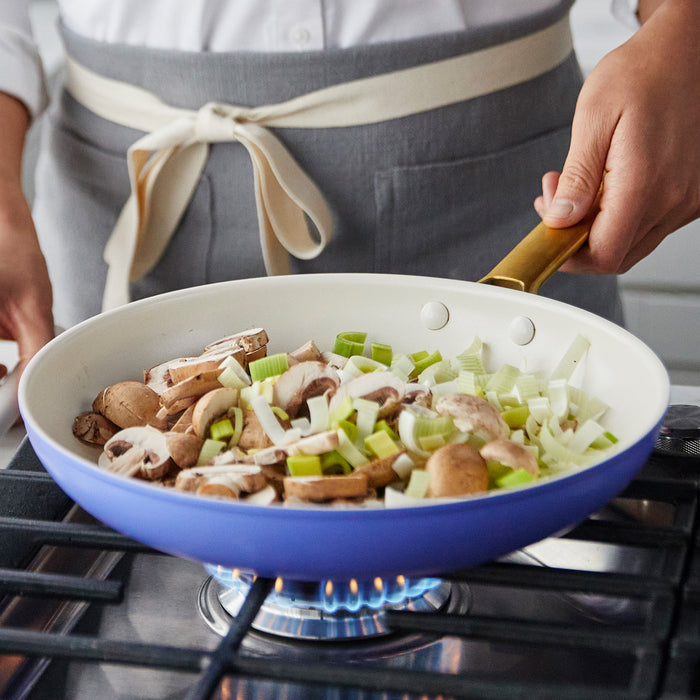 A person in a gray apron cooks mushrooms and celery in a purple Reserve Wisteria frypan on a gas stove.