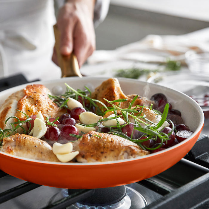 Dark orange frying pan with roasted chicken, grapes, and herbs on a stove.