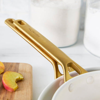 Dark orange frypans with gold handles on a white surface with apples and a cutting board in the background