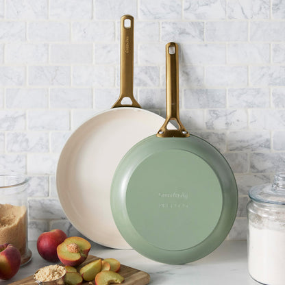 Two green frying pans on a kitchen counter with apples and a cutting board.