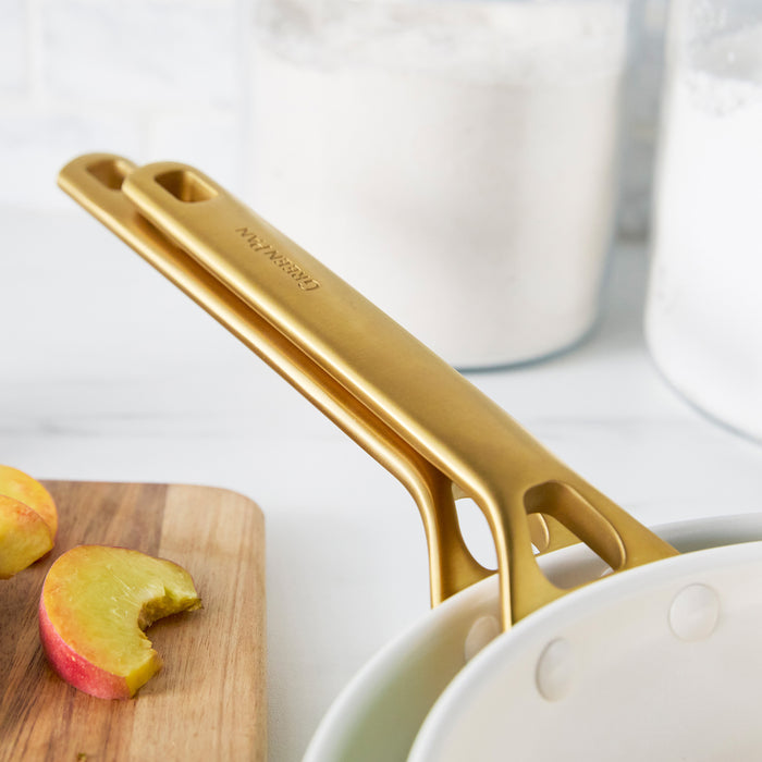 Gold frypan handles on a white surface with apples and a cutting board.