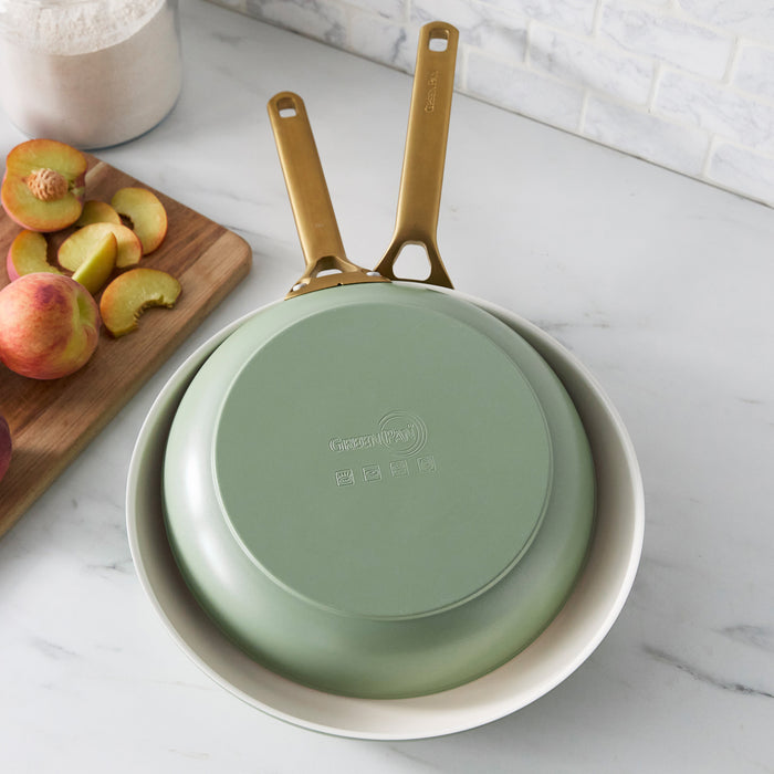 Two green frying pans with gold handles on a marble surface with peaches and a cutting board.