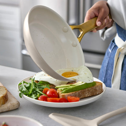 Person using a taupe frying pan with a gold handle to place an egg on a plate with avocado and tomatoes.