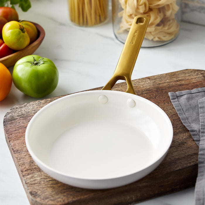 Taupe gray frying pan with gold handle on a wooden cutting board, surrounded by fruits and pasta.