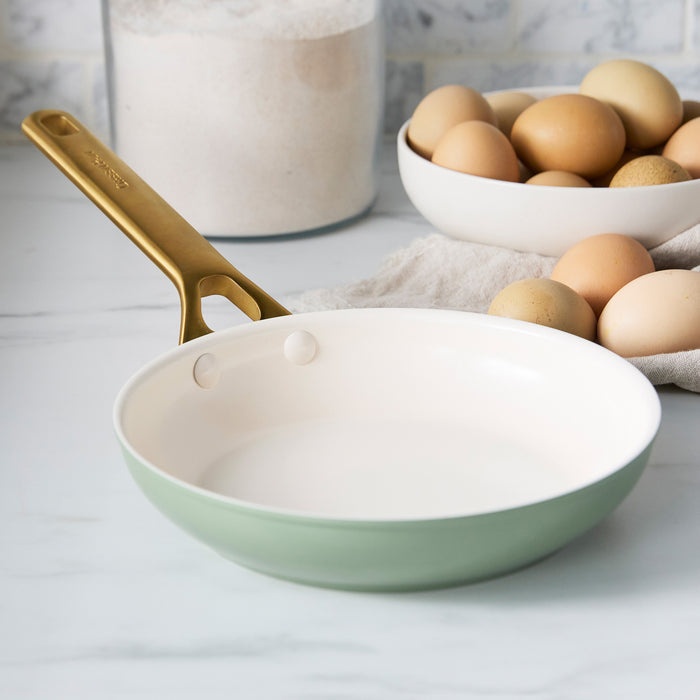 Sage green frying pan with gold handle on a light surface with eggs and flour in the background