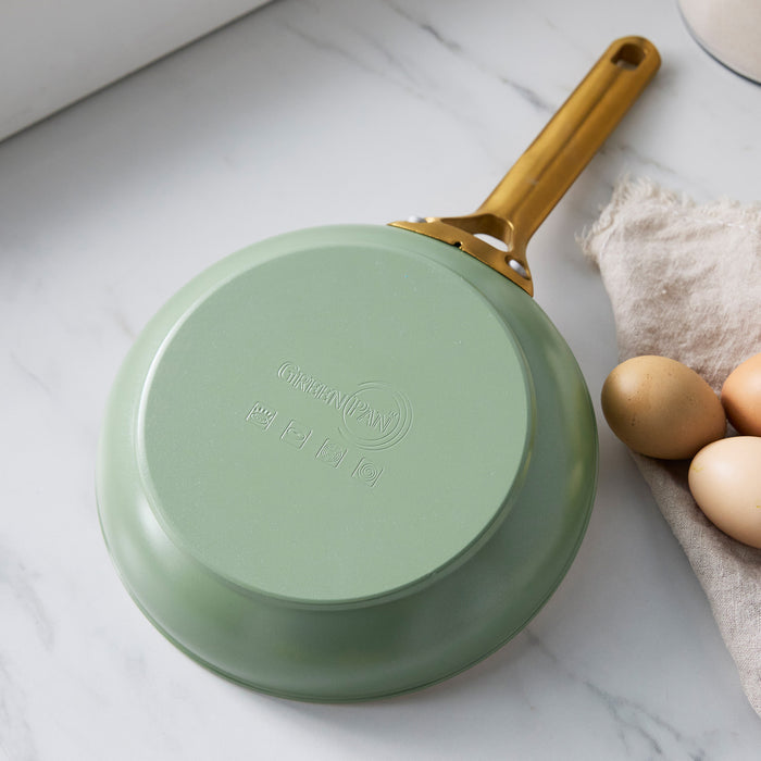 Sage green frying pan with a gold handle on a marble surface with eggs and flour.