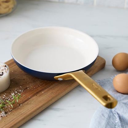 Dark blue frying pan with a gold handle on a kitchen counter with eggs and a cutting board.