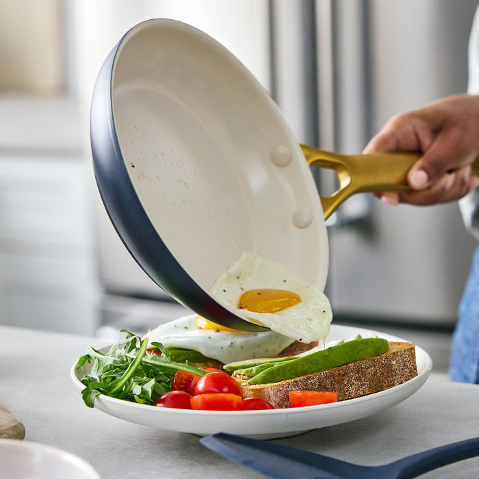 Person holding a dark blue frying pan with eggs over a plate of toast and vegetables