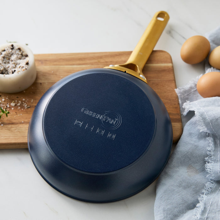 Dark blue frying pan with a gold handle on a wooden cutting board with eggs and a towel in the background.