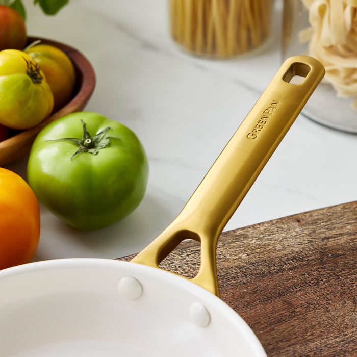 Gold frypan handle on a wooden surface with tomatoes and pasta in the background